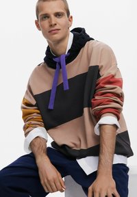 Sudadera en bloques de color en tonos beige, negro, rojo y mostaza, con un cordón morado, puños acanalados y un bolsillo tipo canguro. Textura de tela suave.