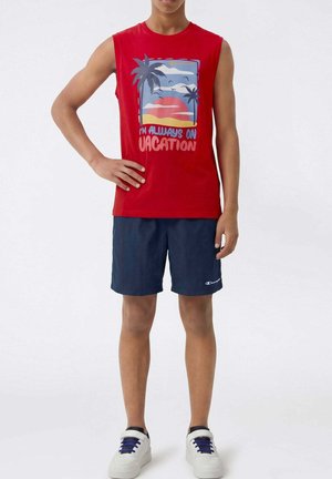 Persona che indossa una maglietta rossa senza maniche con grafica di palma e testo "I'm always on vacation,", pantaloncini blu navy e sneakers bianche, in piedi con una mano sul fianco.