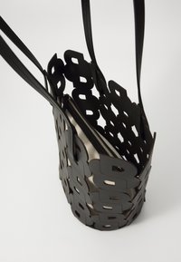 Bolso tote negro con diseño recortado que presenta formas de letras, doble asa y cierre de cremallera. En el interior tiene un forro de tela texturizada.