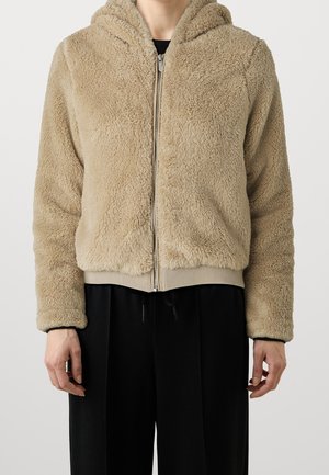 Person iført en beige, blød hættetrøje med lynlås og sorte bukser, stående mod en ensfarvet lys baggrund.