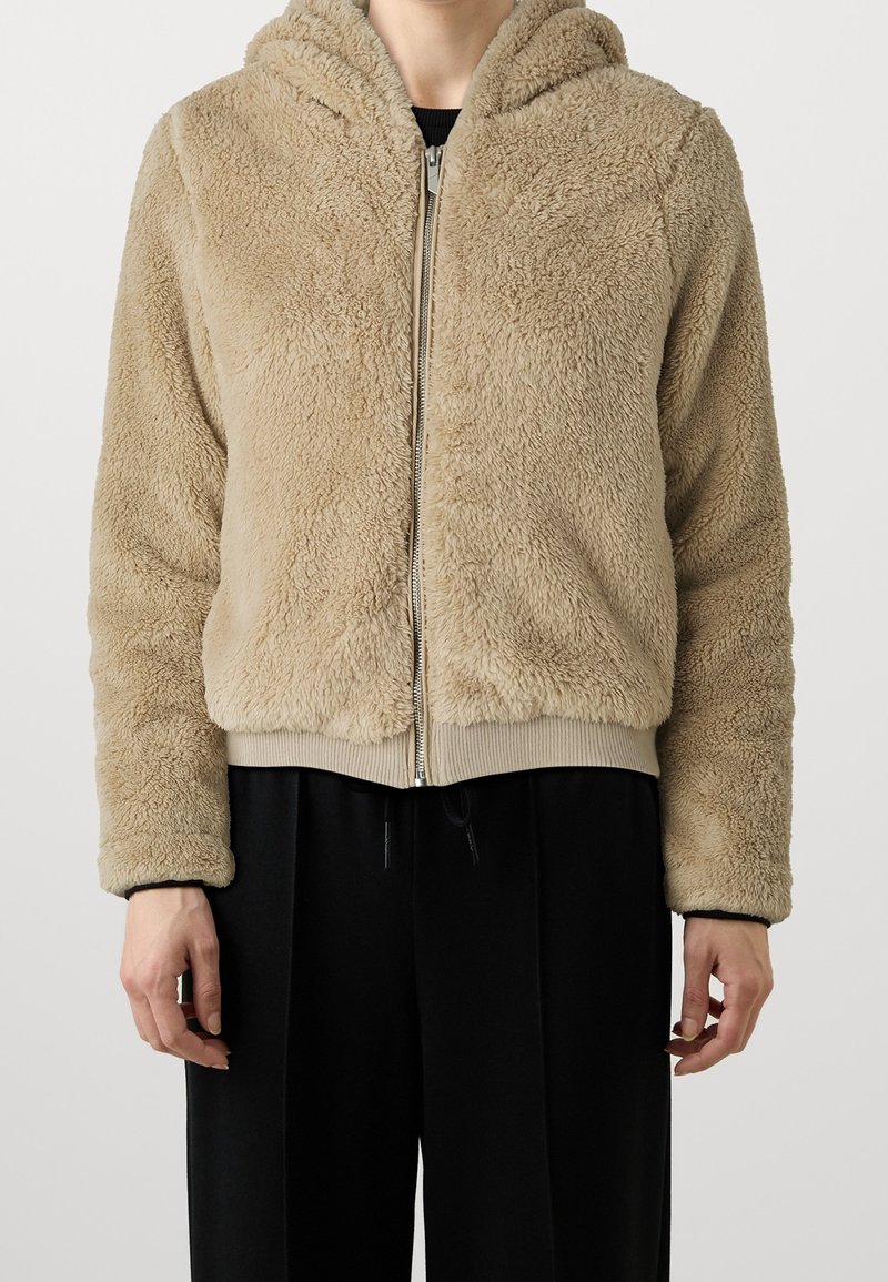 Personne portant un sweat à capuche beige duveteux avec fermeture éclair et un pantalon noir, debout devant un fond clair uni.