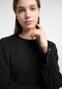 Felpa nera con scollo rotondo, maniche arricciate e dettagli elastici. Texture morbida e vestibilità rilassata. Nessun motivo o hardware visibile.
