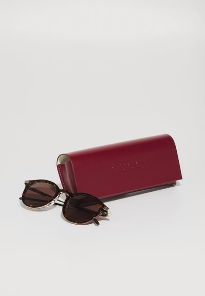 Lunettes de soleil rondes en écaille avec des verres foncés à côté d'un étui en cuir rouge fermé sur une surface blanche.