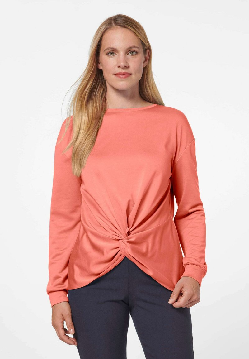 Koraalkleurig langemouwen shirt met een gedraaide voorkant en een ronde halslijn, gemaakt van zachte stof. Gepaard met donkere broeken. Eenvoudige, minimalistische stijl.