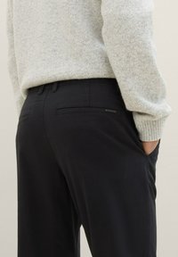 Pantaloni neri con vestibilità su misura, dotati di due tasche posteriori e un discreto etichetta del marchio. Abbinati a un maglione grigio chiaro con texture.