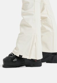 Witte skibroek met een naadloos ontwerp en ritsen bij de manchetten, gecombineerd met zwarte skischoenen met hardwaredetails en gestructureerde zolen.