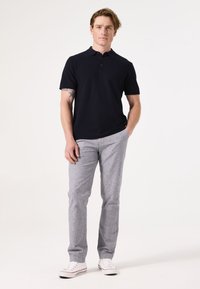 Marinefarbenes Kurzarm-Polohemd mit strukturiertem Muster, kombiniert mit hellgrauen, gemusterten Hosen und weißen Turnschuhen. Das Model steht mit den Händen in den Taschen.