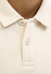 Beige polotröja med klassisk krage, två beige knappar och en texturerad yta. Sömmarna är synliga längs kanterna.