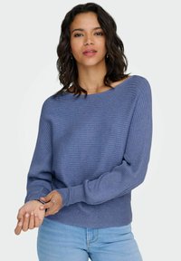 Blauwe geribbelde trui met boothals, lange mouwen en een aansluitende zoom. Draagt over lichte spijkerbroek. De stof is zacht en dik.