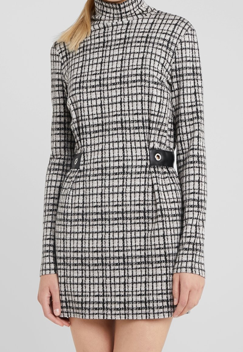 Femme portant une robe mini à manches longues à carreaux noirs et blancs avec un col montant et des détails de boucle noire à la taille.