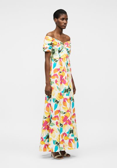 Sieviete valkā garu, no pleciem nolaistu balta kleitu ar krāsainiem, lieliem ziedu rakstiem un bēšām, atvērtām papēžu kurpēm, stāvot pret vienkāršu fonu.