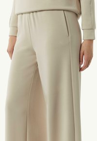 Jasne beżowe spodnie w szerokim kroju z gładką fakturą, z bocznymi kieszeniami i elastycznym paskiem w talii dla większego komfortu. Prosty i nowoczesny design.