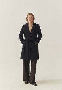 Marineblauer Wollmantel mit maßgeschneidertem Design, mit Schlagkragen, zwei Knöpfen und einem Taillengürtel, kombiniert mit schwarzen weit geschnittenen Hosen.