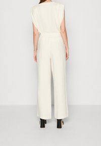Brede jumpsuit van romig beige stof, met subtiele textuur, een elastische taille en kapmouwen. Zwarte block-heeled schoenen eronder.
