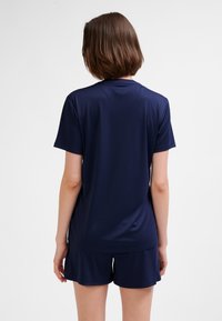 Vrouw met kort bruin haar, gekleed in een effen marineblauw shirt met korte mouwen en bijpassende korte broek, met de rug naar de camera tegen een witte achtergrond.