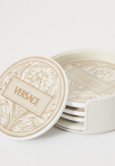 Sada podtácků Versace v kulatém bílém držáku. Podtácky mají květinový zlatý a krémový design s vyraženým nápisem "VERSACE" na střední destičce.