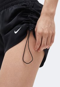 Κοντινό πλάνο στο επάνω μέρος του μηρού ενός ατόμου που φοράει μαύρο σορτς Nike με ριμπ λεπτομέρεια στο πλάι και ρυθμιζόμενο κορδόνι, το χέρι ακουμπά δίπλα στο πόδι.