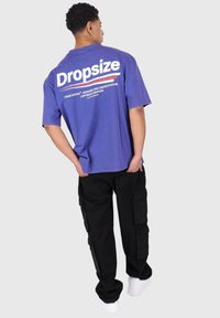 Lila T-Shirt mit weißem "Dropsize"-Text, kurzen Ärmeln und lässiger Passform. Kombiniert mit schwarzen Cargo-Hosen mit seitlichen Taschen.