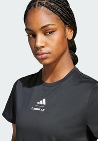 Sort sports t-shirt med korte ærmer, som har en hvid grafik og teksten "LES MILLS" på brystet. Glat stoftekstur.