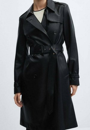 Femme portant un trench-coat en cuir noir à double boutonnage avec ceinture, poches à rabat et épaulettes boutonnées, par-dessus un haut blanc.