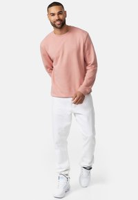 Sudadera rosa clara con cuello redondo, puños acanalados y un pequeño logo en el pecho, combinada con pantalones vaqueros blancos y zapatillas blancas.