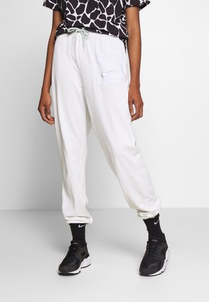 Osoba nosząca białe spodnie dresowe Nike ze sznurkiem, czarne skarpetki Nike, czarne buty sportowe oraz czarno-biały wzorzysty top krótko powyżej talii.