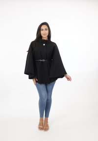 Svart cape-liknande topp med hög krage, knäppning fram och ett svart bälte som accent, kombinerad med ljusblå slim fit jeans och genomskinliga klackar.