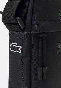 Borsa in tessuto nero con tracolla nera, zip con tirazip brandizzati "Lacoste" e logo del coccodrillo ricamato in nero, bianco e rosso.
