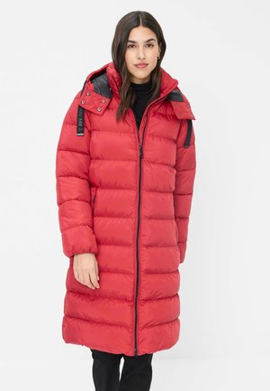 Frau trägt einen langen roten gesteppte Mantel mit schwarzem Reißverschluss und Kapuze, steht vor einem schlichten weißen Hintergrund.