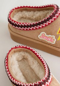 Tan slip-on shoes with a soft, fluffy interior. Red and black braided trim accents the opening. Features "Smile" embroidery and yellow design.