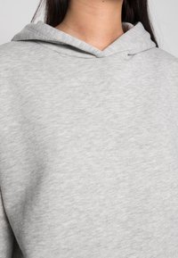 Sudadera gris claro con cordón, hecha de una tela suave y texturizada. Presenta un corte holgado y costuras visibles alrededor de la capucha.