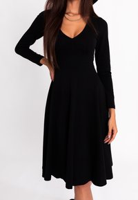 Femme portant une robe noire à manches longues arrivant au genou avec un décolleté en V et deux colliers en chaîne dorée superposés, se tenant avec une main sur la hanche.