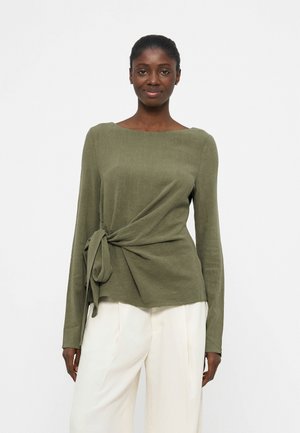 Žena oblečená v olivovo zelenom top s dlhým rukávom a bočným zaväzovaním a biele široké nohavice, stojaca pred jednoduchým bielym pozadím.