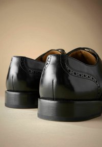 Een paar gepolijste zwarte leren nette schoenen met brogue-details, gezien van achteren op een neutrale beige ondergrond.