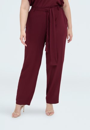 Femme portant un pantalon bordeaux taille haute à jambes larges avec une ceinture nouée et des sandales à talons hauts beiges à brides, debout sur fond blanc.
