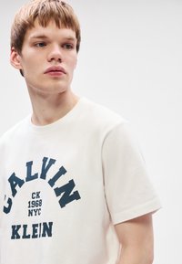 Valge puuvillane T-särk, millel on julge tumesinine graafiline logo, mis ütleb "CALVIN KLEIN 1968 NYC." Klassikaline ümar kaelus ja lühikesed varrukad.