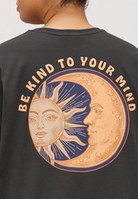 Schwarzes T-Shirt mit einem großen Grafikdesign von Sonne und Mond sowie dem Text "SEI NETT ZU DEINEM GEIST" in Pfirsichfarbe, der sich um das Design windet.