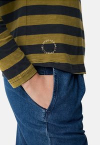 Langärmeliges gestreiftes Hemd in Olivgrün und Schwarz mit rundem Logodetail; getragen mit blauer Jeanshose, Tasche sichtbar.