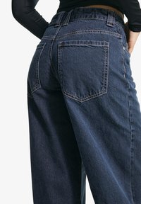 Tmavě modré džíny z denimu s uvolněným střihem, zdobené prošitím, zadními kapsami a mírně zúženým střihem nohavic.