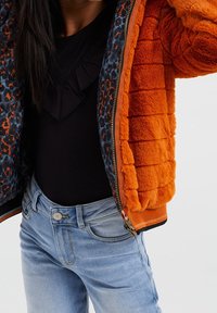 Orange imitationpälsjacka med en kviltad textur, med ett foder i blå och orange leopardmönster. Tillsammans med ljusblå denimjeans.