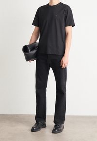 T-shirt nera in cotone con maniche corte e logo discreto, abbinata a jeans neri in denim e scarpe nere, mentre tiene una borsa nera in pelle.