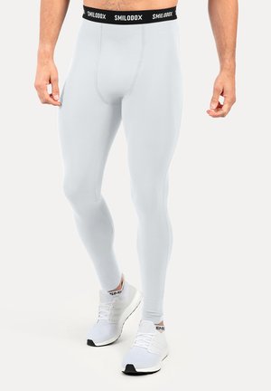 Męski model noszący jasnoszare, dopasowane legginsy sportowe z czarnym pasem oznaczonym "SMILODOX" i białe buty do biegania na białym tle.