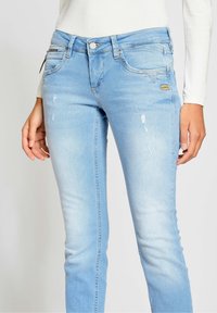 Jeans de denim azul claro con corte slim, que presentan un ligero desgaste, cierre de botón metálico y un pequeño parche de marca en la parte frontal.