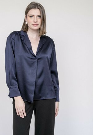 Chemisier en satin navy avec décolleté en V, manches longues et boutons aux poignets ; présente une coupe ample et une texture lisse. Associé à un pantalon noir.