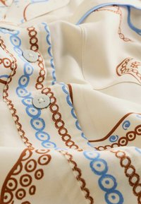 Bluza din bumbac cu un fundal crem, prezentând modele circulare complexe în nuanțe de maro și albastru, și nasturi de un albastru deschis.