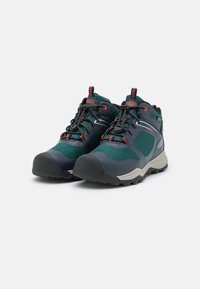 Trail Schuhe mit einem dunkelblauen und grünen Mesh-Obermaterial, ausgestattet mit einer Gummispitze, grauer Sohle und orangefarbenen Akzenten an Schnürsenkeln und Logo.