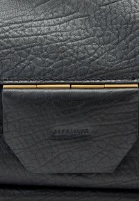 Μαύρη τσάντα από υφή δερμάτινου με εμφανές κούμπωμα από χρυσό μέταλλο. Διαθέτει ανάγλυφο όνομα μάρκας σε μαύρη δερμάτινη λεπτομέρεια.