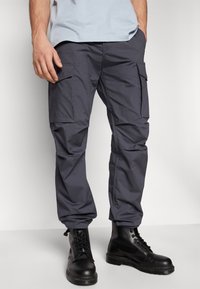 Pantalons cargo gris avec plusieurs poches latérales, fabriqués en tissu léger. Associés à des bottes noires à lacets, cela donne un look robuste.