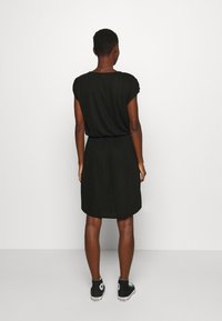 Schwarzes, kurzärmeliges Kleid mit elastischem Bund. Der Stoff ist weich, und der Saum endet knapp über den Knien. Kombiniert mit schwarzen Sneakern.
