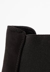 Bottine noire avec une tige en daim, un panneau en tissu texturé, des coutures renforcées et un bord lisse à l'ouverture.